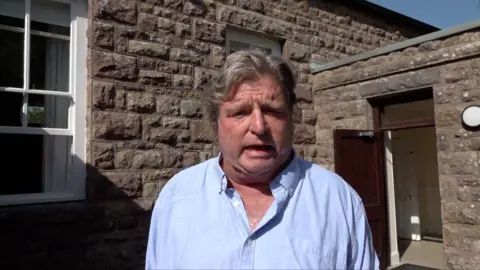 A man in a light blue top with grey hair. He is stood in front of a brown building. The sky is blle.