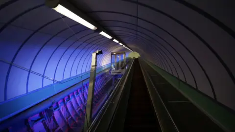 A glass lift travelling along an incline. It has a steel framework. The tunnel area is illuminated.