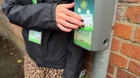 The hand of an unidentifiable person pressing a blue and green credit card-sized card to a grey box attached to a lamppost with a brick wall behind it.