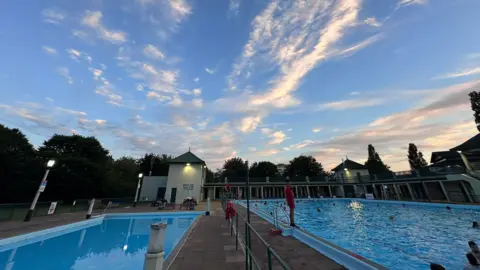 Image of Peterborough lido, with several swimmers in the pool and a lifeguard wearing red watching over. The sky ahead is blue.