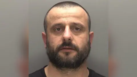 A police mugshot of Florjan Ceka. He has very short black hair and a black moustache and beard. He is wearing a black top and there is a grey background.