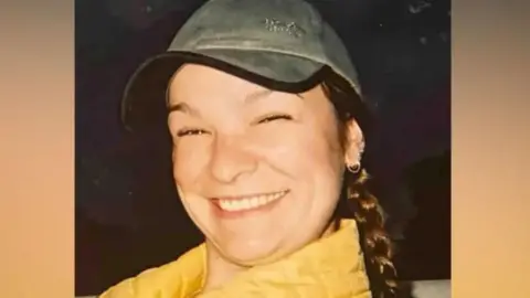 Clare smiles widely at the camera. She is wearing a green and black cap and a yellow puffer jacker, and has lots of earrings in one ear. Her hair is in a long dark plait. 