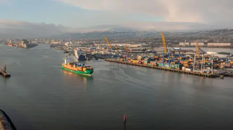 Belfast Harbour A ship pictured sailing with Belfast port in the background. The ship is green carrying yellow shipping containers.