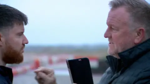 Barry Elliott has short greying hair, combed forwards from the temples, with a slight peak at the forehead and a grizzled white grey beard and slight moustache. He is wearing a black coat with the collar up. He is mid sentence, with his mouth open, looks stern and is pointing a finger at a BBC reporter. He has his mobile phone in the other hand.