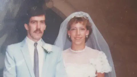 Linda Norton A married couple in the 1980s. The groom is wearing a pale blue suit, white rosette and moustache. The bride has blonde hair and is wearing a white dress with a veil.