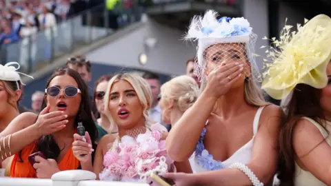 Reuters Three women, one in an orange dress and another wearing a dress covered in pink flowers, cover their mouths and gasp as they watch the racing 