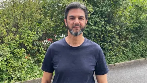 Sofia Luis-Hobbs/BBC A person wearing a dark short-sleeved T-shirt stands outdoors in front of dense green shrubs and trees. The image is a waist-up portrait taken on a paved path, with foliage filling most of the background.