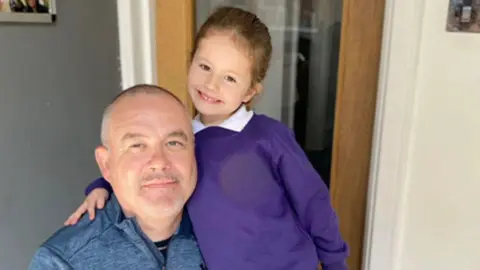 Connie pictured at a young age with her dad Paul. She is dressed in school uniform and her dad is wearing a blue zip sports top. He has cropped greying hair and stubble.