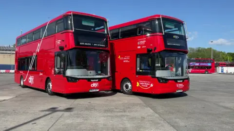Two red double-decker buses are parked side by side in an open area. Both display the text '100% electric • zero emissions' on the front. The left bus has 'city' written on it, while the right bus includes branding for a project with Cornwall Council and partners. Additional red buses are visible in the background.

