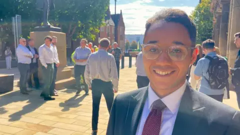 BBC A man in glasses, a dark purple tie, white shirt and black suit stands close to a statue. A dozen men stand casually behind him, some are wearing shirts and trousers, others are wearing luminous workwear.