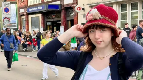 Handout/PA RealLife Amelie Rule standing in the street in front of Camden Town tube station. She is posing for the camera with her hands holding a red knitted hat, and she is wearing a blue cardigan and white t-shirt