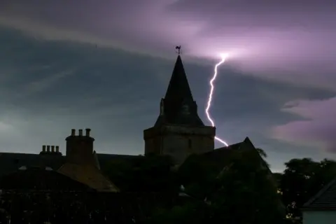 Zara Macdonald Dornoch Cathedral under the night sky. It is dark and there is a lightning bolt in the purple-coloured sky.