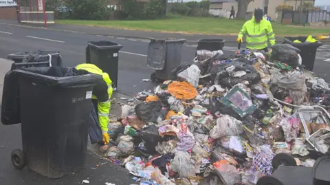 Two men in high vis jackets working on a large pile of rubbish, which has been emptied on to a road. Black wheelie bins surround the pile. In the background on the other side of the road is a bus stop and a pedestrian in the distance.