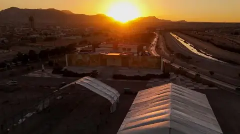 A giant tent, with white material, in the foreground. Behind it is a semi rural arid landscape. In the distance the sun pokes over the hills in the distance. 