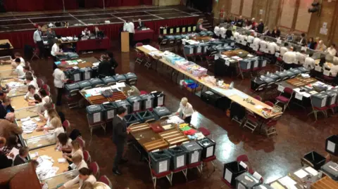 An aerial view of a ballot counting hall. People sit at tables counting ballots. 