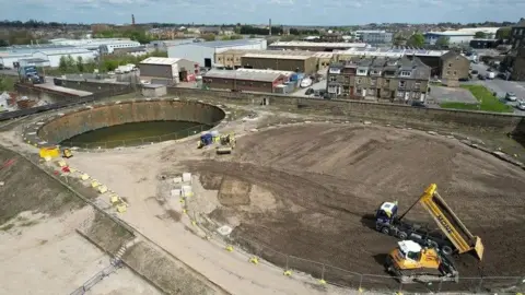 Diggers sit on what appears to be an aerial view of a building site, which is covered over with sand. An industrial-sized well of water sits in the corner