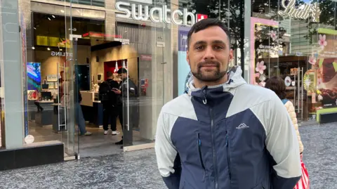 A man, in his late 20s or early 30s, with black hair and a black beard, smiles at the camera outside a glass-fronted shop with the Swatch logo above the door in illuminated white lettering. He is wearing a grey and blue rain coat. 