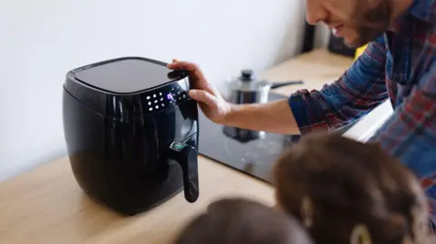 A man in a blue and red chequered shirt is pressing a button on a black air fryer on the table. Two child's heads can be seen stood next to him.