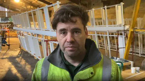 Luke Deal/BBC Timothy Whiting looks at the camera in a warehouse. White windmill sails rest behind him in the warehouse. He has brown hair that overhangs on his forehead and he is wearing a florescent green coat. 