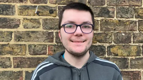 Callum has short dark brown hair, black-frame glasses and is wearing a grey Adidas-branded hooded top. There is a brick wall behind him.