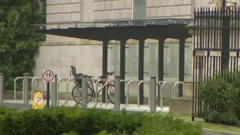 A metal and glass bike shelter with metals railings to prop bicycles against. One bike is in the shelter. Behind the shelter a grey building