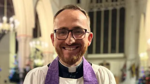 Tom Mumford smiles at the camera inside Ipswich Minster. He has short brown hair, glasses, a thick beard and he wears a vicar collar and robe.