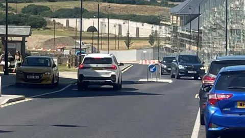 Several cars in the foreground of a wide shot of a road through Nansledan. There is a white road bridge, and countryside fields and trees with a blue sky in the background. The road has houses being built with scaffolding on the right hand side  and shops on the left.