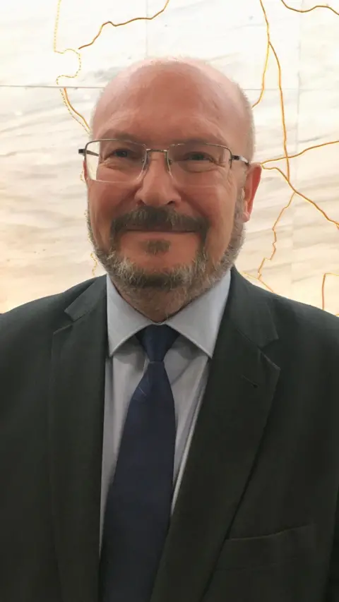 Graham Plant wearing a grey suit and a blue tie. He is bald, has a beard and wears silver-framed glasses