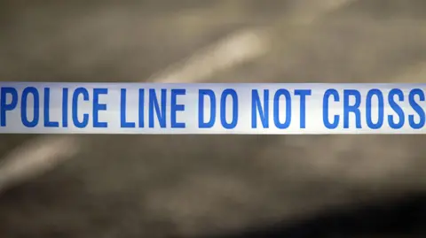 Peter Byrne/BBC A blue and white line of tape is stretched across a road. it has the words police line do not cross om it in blue.