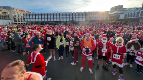 Hundreds of runners dressed in Santa costumes stand ready to run in the middle of a square. 