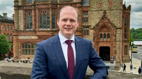 dfc a man in a blue suit, wear a white shirt and crimson tie. he is standing , with clasped hands, on Derry's walls overlooking the city's guildhall