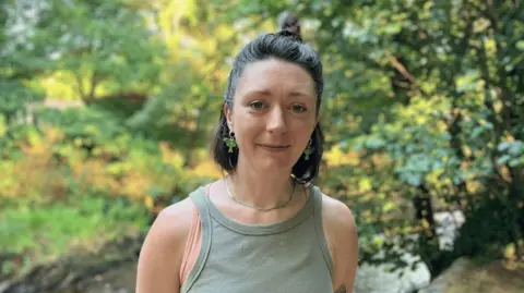 A woman with brown hair and a green vest stands in front of the River Avon at Avonwick, South Devon, where rover levels have fallen after the prolonged dry, hot weather