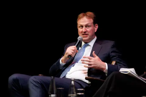 PA Media Jim O'Callaghan sitting on a chair with a microphone. He has brown hair and is wearing a dark suit with a blue tie