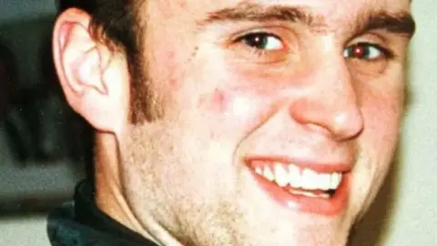 PA Media A man with short brown hair smiling as he looks over his right shoulder at the camera