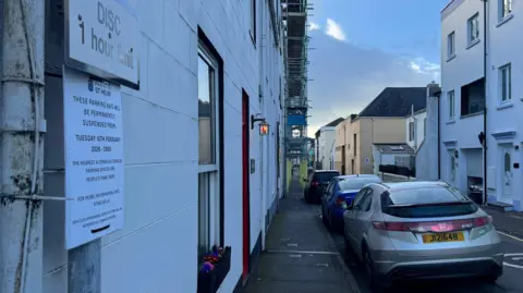 Parking bays on a road in St Helier which are being suspended. Partially cloudy skies with cars in the spaces.
