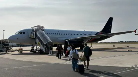 A Titan aircraft, with passengers going up a glazed stairway. It's a stubby jet aircraft with a white body and a blue wing and tail. 