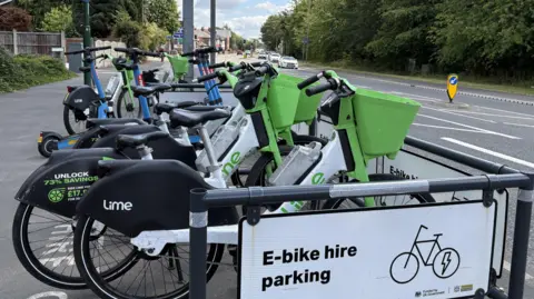 A number of e-bikes and e-scooters parked behind a barrier by the side of a busy road