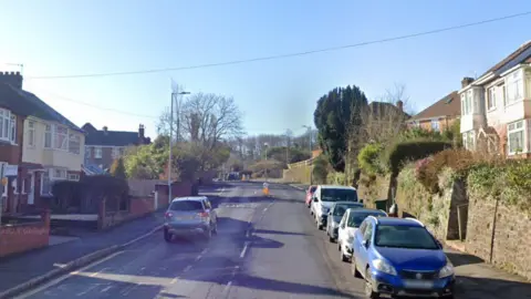 A Google Streetview image of Cowick lane, showing a suburban street with parked cars lining one side. 