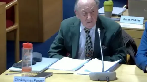 LDRS A man at a council meeting. He has a suit jacket, white shirt and tie on and a water bottle and his name tag 'Cllr Stephen Foster" in front of him. Behind him is the seat of another councillor which has a cup on and the name "Sarah Wilkins". 