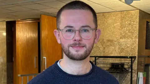 Connor wears a dark blue jumper and has round glasses and tightly shaved dark hair and beard, He is standing inside county hall with doors and wood panelled walls behind them and some railings with coat hangers. 