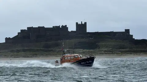 RNLI/Tom Collins The blue and orange RNLI lifeboat in the water in front of Bamburgh Castle.