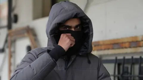 PA Media Imran Chowdhury outside court wearing a coat with the hood up and covering his face with a black material so only his eyes can be seen.