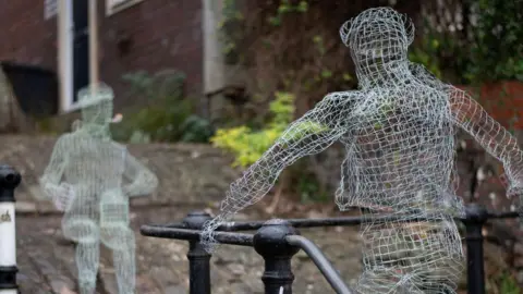 Jo Bushell/Barnaby Johnson Two figures made from wire mesh face each other on the cobbles of St Luke's steps in Totterdown, Bristol. One of them is standing holding the rail alongside the stairs and another is sitting down smoking a pipe