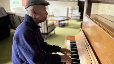 Mr Stephenson is sat playing a light-brown piano. It is inside his home.