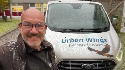 Andy Crowle Andy Crowle smiling for the camera in front of the Urban Wings white company van. There is a hawk drawn on the bonnet. It is snowing. There is a building behind him.