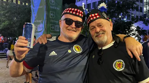 David Grindlay and Alistair Fleming smiling for the camera in Boston. David is holding up a drink can and is wearing sunglasses. Bot men are wearing blue Scotland tops and tam o'shanters.