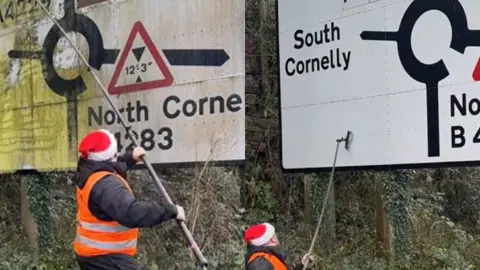 Clean Cymru Two images of a man cleaning a road sign. He is wearing an orange hi-vis jacket and a Santa hat. The image on the left the road sign is dirty, and the one on the right is clean and it can be read. The road sign shows Porthcawl, South Cornelly and North Cornelly. It is a roundabout road sign.