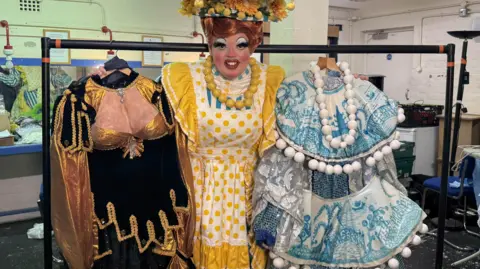 A man wearing a yellow, white and blue dress with polk apron smiling with lots of make up and a floral hat on. He's holding two other pantomime dame dresses. 