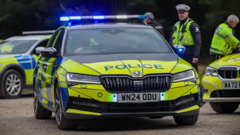 A black, yellow and blue Skoda police car from Devon and Cornwall Police drives off with its blue lights on. Three police officers are stood near the car and two other police vehicles which are parked up.