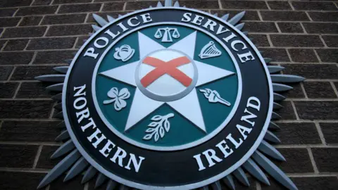 Getty Images A PSNI crest. The outer circle is black, the inner circle is green and in the center is a star with St. Patrick's Saltire as well as a shamrock, torch, crown, harp and scales. 